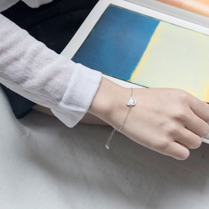 Puede incluir: Un delicado brazalete de cadena de plata con un dije en forma de corazón blanco. El brazalete está en la muñeca de una persona.