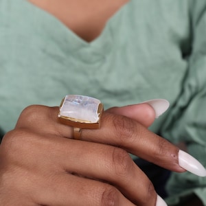 Peut inclure: Une bague dorée ornée d'une grande pierre de lune de coupe carrée. La pierre de lune a un aspect blanc laiteux et irisé. La bague est sertie dans un métal doré avec un anneau simple. C'est un bijou.
