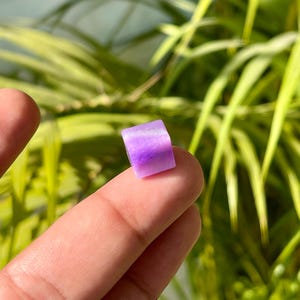 May include: A small, square, purple gemstone held between fingers. The gemstone has a gradient of colors, from a deep purple to a lighter lavender hue. The background features green foliage.