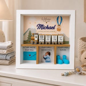 Könnte beinhalten: Eine weiß gerahmte Baby-Andenkenbox mit dem Namen "Michael" in blauer Schrift. Die Box enthält einen Zug, Uhren und einen Heißluftballon. Der Text enthält "A TINY NOTE FOR YOU", "OUR FIRST MEMORIES" und "YOUR FIRST SOCKS".