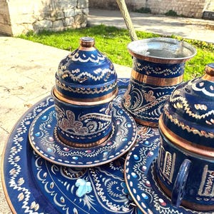 May include: A traditional Turkish coffee set featuring a dark blue tray, cups, and saucers with intricate copper and silver-toned designs. The set includes a coffee pot and a serving spoon, all with matching patterns. The set is designed for serving Turkish coffee.