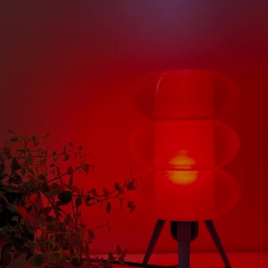 Può includere: Una lampada da tavolo rossa in stile retrò con un design a paralume ovale impilato. La lampada ha una base a treppiede grigia ed emette un bagliore caldo. Una piccola pianta con foglie verdi è visibile in primo piano.