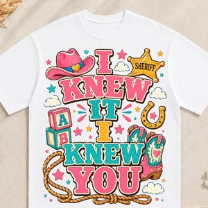 May include: A white t-shirt featuring a colourful graphic print with the text I KNEW IT I KNEW YOU. The design includes a pink cowboy hat, a yellow sheriff star badge, alphabet blocks, a horseshoe, a lasso, and pink cowboy boots with hearts.