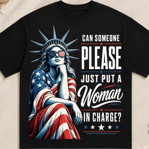 May include: A black t-shirt featuring a graphic of the Statue of Liberty wearing American flag sunglasses. The text reads: CAN SOMEONE PLEASE JUST PUT A Woman IN CHARGE? The design includes red, white, and blue accents with stars and stripes motifs.