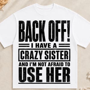 May include: A white short-sleeved t-shirt featuring black distressed-style text that reads: BACK OFF! I HAVE A CRAZY SISTER AND I'M NOT AFRAID TO USE HER. The graphic is centered on the chest and uses a bold, block-style font with horizontal lines.