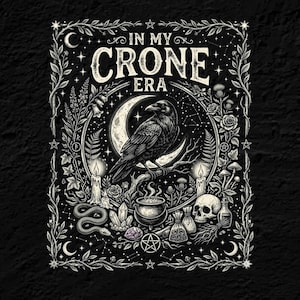 May include: A black and white graphic print featuring a raven perched on a crescent moon, surrounded by candles, a cauldron, a skull, crystals, and botanical elements. The text IN MY CRONE ERA is centered at the top within a decorative floral border.