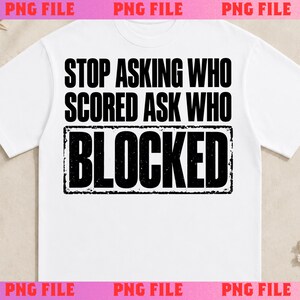 May include: A white short-sleeved t-shirt featuring black text that reads STOP ASKING WHO SCORED ASK WHO BLOCKED. The word BLOCKED is enclosed in a black rectangular border with a distressed, textured appearance.