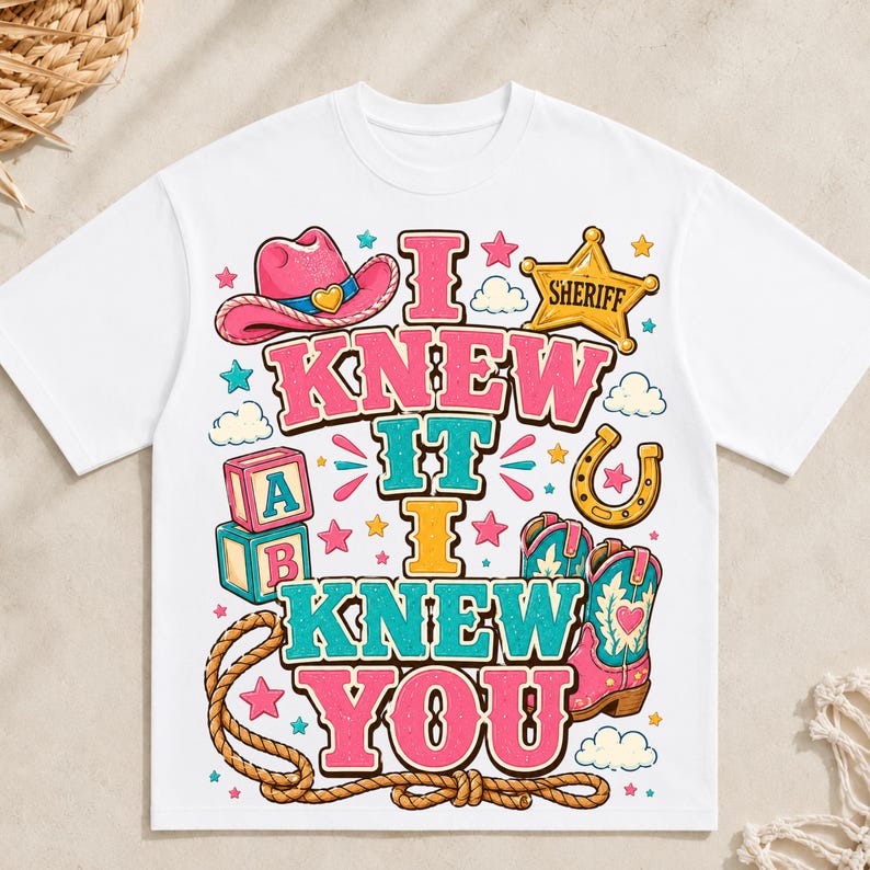May include: A white t-shirt featuring a colourful graphic print with the text I KNEW IT I KNEW YOU. The design includes a pink cowboy hat, a yellow sheriff star badge, alphabet blocks, a horseshoe, a lasso, and pink cowboy boots with hearts.