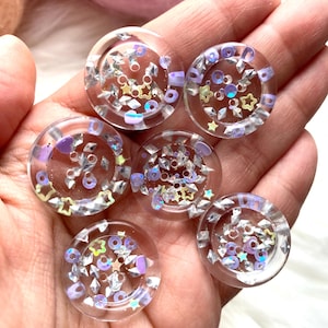 May include: Six clear, round buttons with iridescent glitter and star-shaped accents. The buttons have a flat back and are filled with a mix of silver, purple, and gold glitter. The buttons are held in a hand.