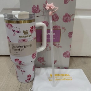 May include: A white Stanley Quencher H2.0 tumbler with a floral rose pattern, a white handle, and a gold rim. Includes a pink bow-topped straw, a white drawstring bag, and a matching floral box. The tumbler is 40 oz.