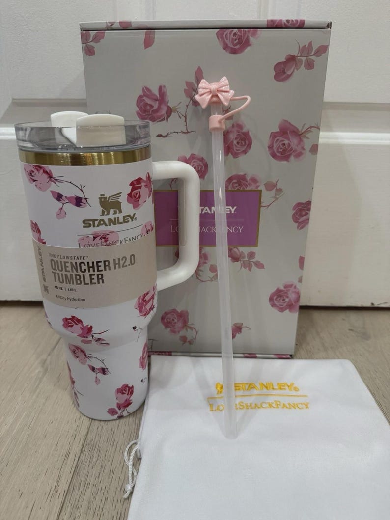 May include: A white Stanley Quencher H2.0 tumbler with a floral rose pattern, a white handle, and a gold rim. Includes a pink bow-topped straw, a white drawstring bag, and a matching floral box. The tumbler is 40 oz.