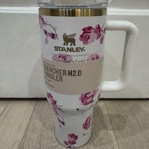 May include: A white Stanley Quencher H2.0 tumbler with a floral rose pattern in pink and purple. The tumbler has a white handle, a clear lid, and gold accents. The text "STANLEY" and "LOVESHACKFANCY" are visible.