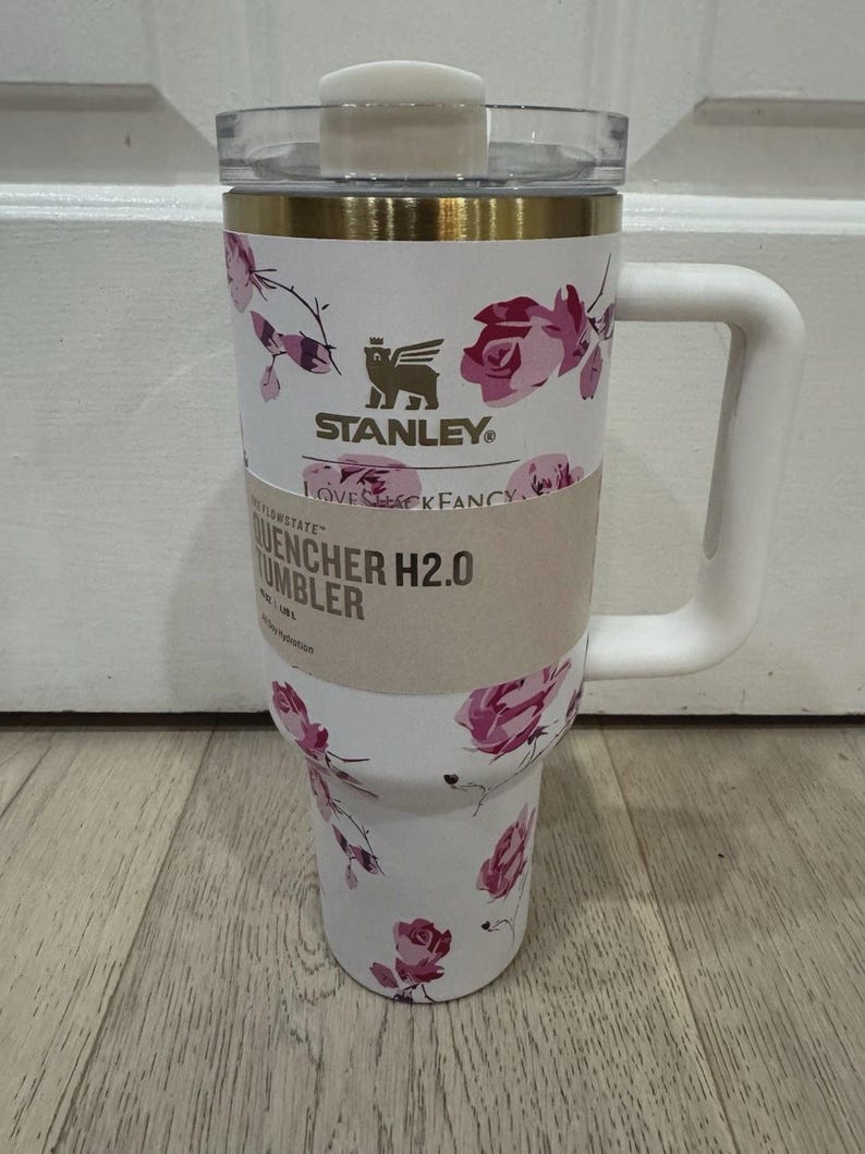 May include: A white Stanley Quencher H2.0 tumbler with a floral rose pattern in pink and purple. The tumbler has a white handle, a clear lid, and gold accents. The text "STANLEY" and "LOVESHACKFANCY" are visible.