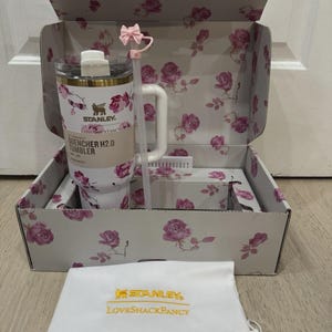 May include: A white Stanley Quencher H2.0 tumbler with a floral rose design, inside a matching box. The box and tumbler feature pink roses and green leaves. A white bag with gold text and a pink bow straw complete the set.
