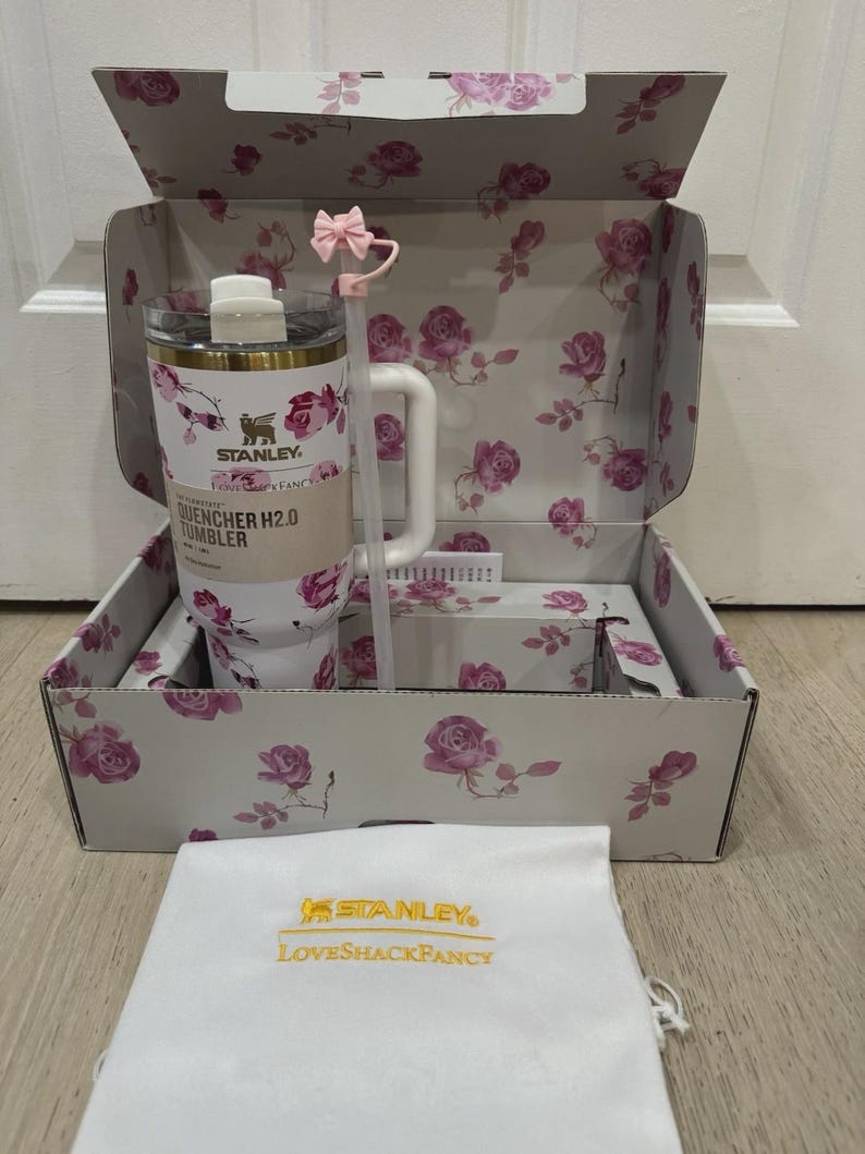May include: A white Stanley Quencher H2.0 tumbler with a floral rose design, inside a matching box. The box and tumbler feature pink roses and green leaves. A white bag with gold text and a pink bow straw complete the set.