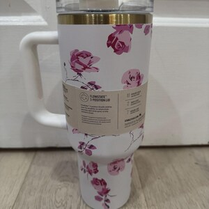 May include: White insulated tumbler with a handle, featuring a floral rose pattern in shades of pink. The tumbler has a clear lid and a gold band near the top. A tag indicates a 3-position lid.