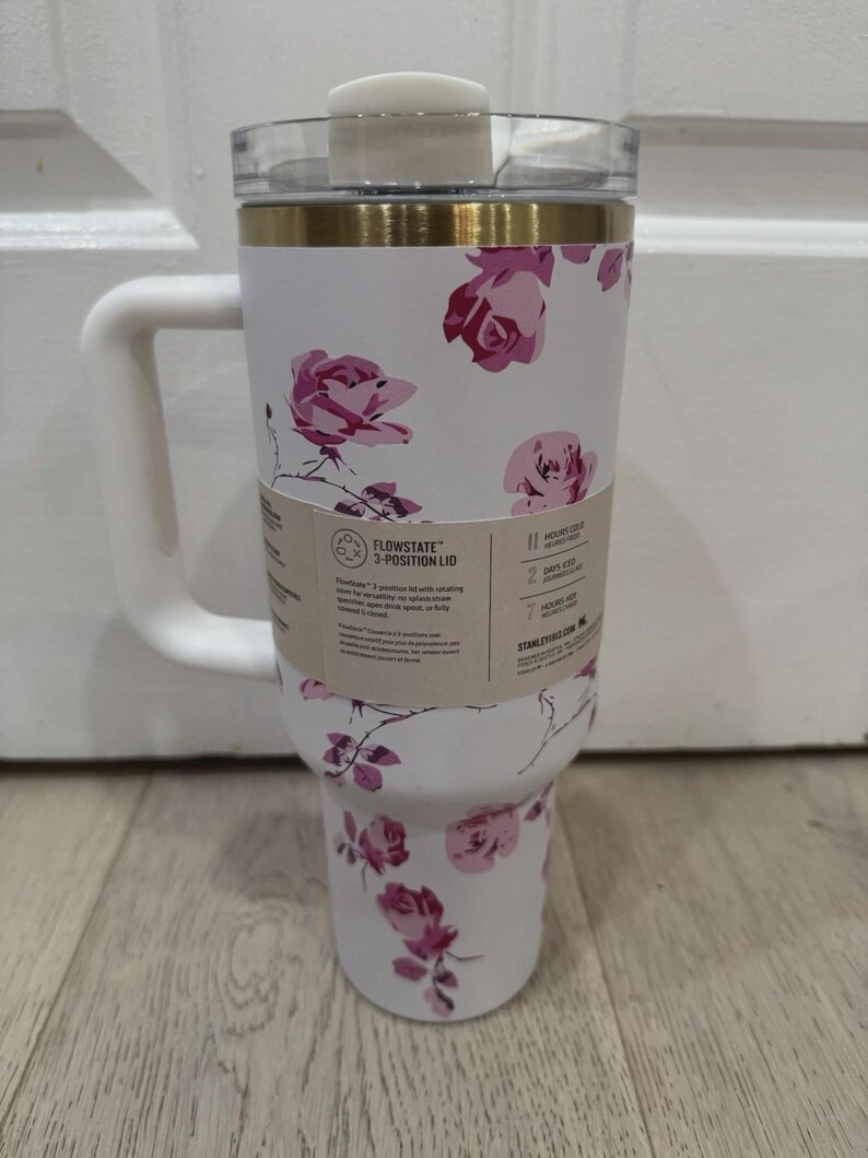 May include: White insulated tumbler with a handle, featuring a floral rose pattern in shades of pink. The tumbler has a clear lid and a gold band near the top. A tag indicates a 3-position lid.