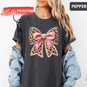 May include: Dark gray t-shirt featuring a large bow design. The bow has a leopard print pattern with a pink center and a decorative border. The text "FLAT 2D PRINTING" is visible on the upper left. The word "PEPPER" is in the upper right.