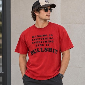 May include: A red short-sleeved t-shirt featuring black text that reads DANCING IS EVERYTHING EVERYTHING ELSE IS BULLSHIT. The shirt is paired with dark gray trousers, a black and tan baseball cap, and dark sunglasses with black frames.