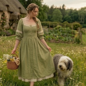 Puede incluir: Una mujer con un vestido verde salvia con detalles de encaje lleva una cesta de mimbre llena de flores, caminando por un campo de flores silvestres con un perro. El vestido tiene botones y una enagua blanca. Una cabaña con techo de paja está al fondo.