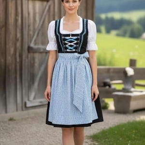 以下が含まれることがあります： 伝統的なバイエルン風のディアンドルドレスを着た女性。ドレスは、青いトリムが付いた黒い胴着と白いブラウスが特徴です。青と白のストライプのエプロンはウエストに結ばれています。彼女は黒い靴と白い靴下を履いています。背景には木造の建物と緑の風景があります。