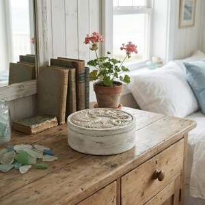 May include: A weathered wooden table displays a round, white decorative box with a carved design, alongside vintage books and a potted plant with pink flowers. A jar of sea glass and a mirror are also visible.