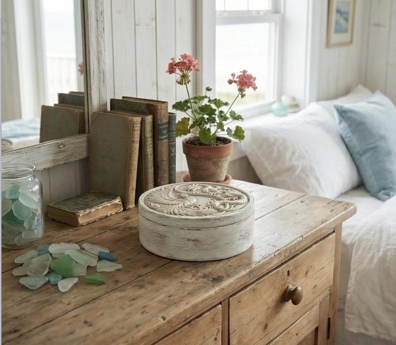 May include: A weathered wooden table displays a round, white decorative box with a carved design, alongside vintage books and a potted plant with pink flowers. A jar of sea glass and a mirror are also visible.