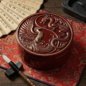 May include: A round, red container with a detailed relief carving of a dragon and phoenix in a yin-yang design. The container sits on a red patterned cloth, with a calligraphy brush and fan in the background.