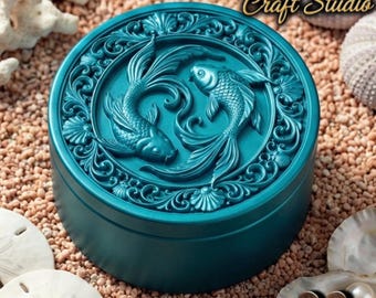 May include: Teal-colored round box with a detailed relief of two koi fish. The box is surrounded by decorative scrollwork and is labeled "Design by Fortuna Craft Studio". The box is sitting on a bed of sand with seashells.