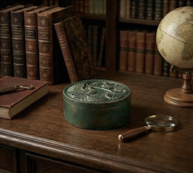 May include: A round, antique-style green box with a detailed bird design on the lid. The box sits on a wooden surface, alongside a magnifying glass and a globe. Bookshelves filled with old books are in the background.