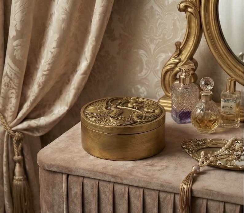 May include: A gold-colored round box with an ornate phoenix design on the lid sits on a velvet-covered surface. The box is next to perfume bottles and a decorative mirror. The background includes a patterned curtain.