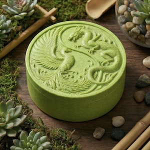 May include: A round, light green box with a detailed relief carving of a dragon and phoenix in a yin-yang design. The box is surrounded by succulents and moss, creating a natural aesthetic. The box is likely used for storage or display.