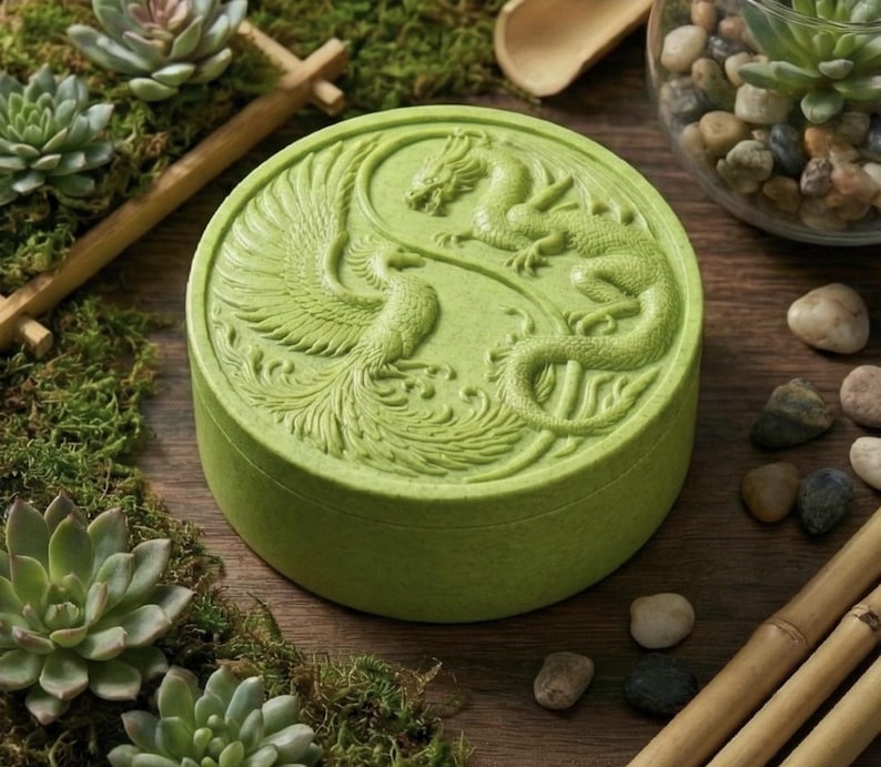 May include: A round, light green box with a detailed relief carving of a dragon and phoenix in a yin-yang design. The box is surrounded by succulents and moss, creating a natural aesthetic. The box is likely used for storage or display.