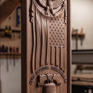 May include: A rectangular dark wood plaque featuring a relief carving of an eagle perched atop a draped American flag. Below, a Liberty Bell is carved with the text FAITH FREEDOM INDEPENDENCE, 250 YEARS, and the dates 1776-2026 at the bottom.