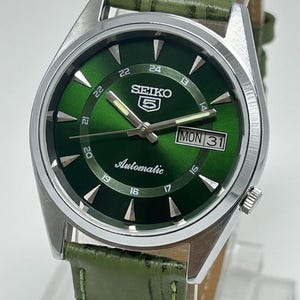 Puede incluir: Un reloj automático Seiko 5 con esfera verde y correa de cuero verde. El reloj tiene una caja plateada y muestra el día y la fecha. La esfera muestra el logotipo de Seiko 5 y la palabra "Automatic".