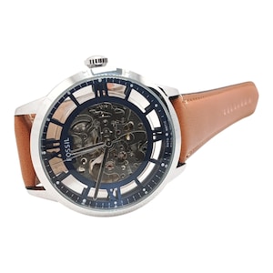 Puede incluir: Un reloj Fossil plateado con una correa de cuero marrón. La esfera del reloj tiene un centro transparente, que revela el intrincado mecanismo interno. El reloj presenta números romanos y la palabra "Fossil" en la esfera.
