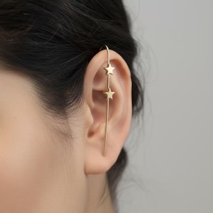 May include: A gold-coloured ear cuff with two star charms. The ear cuff curves over the top of the ear and extends down with the star charms suspended along the length of the ear. The jewellery is worn on the ear.