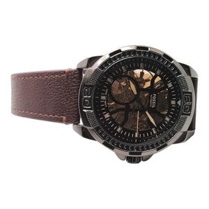 May include: A Fossil automatic watch with a black face and a brown leather band. The watch has a visible inner mechanism and a silver-tone bezel. The band is a medium brown color.