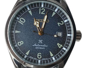 May include: A Seiko automatic wristwatch with a textured dark blue dial, silver-tone Arabic numerals, and luminous hands. The watch features a date window at the three o'clock position, a 20 BAR water resistance rating, and a polished silver-tone case.