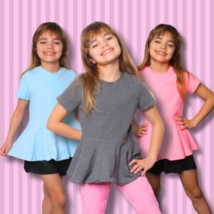 May include: Three young girls wearing short-sleeved tops with flared peplum hems. One girl wears a light blue top, another a gray top, and the third a pink top. They are also wearing black shorts and pink leggings. The background is a pink striped pattern.