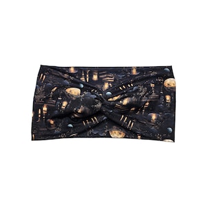 May include: Black and gold headband with a celestial print. The fabric features a repeating pattern of planets, buildings, and glowing lights. The headband is knotted at the top.
