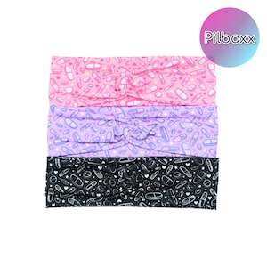 May include: Three headbands in pink, purple, and black, each patterned with white pill and heart shapes. The pink headband has a light pink background, the purple has a lavender background, and the black has a black background. The brand name "Pilboxx" is in a circle in the upper right corner.