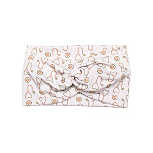 May include: A light gray headband with a twist design. The headband is patterned with orange stethoscopes, hearts, and medical crosses. The fabric is soft and appears to be made of a stretchy material.
