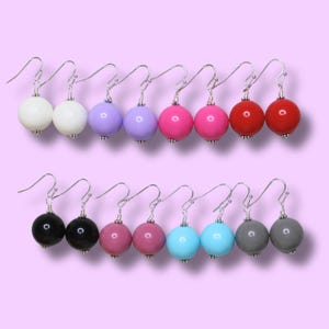 May include: A collection of colorful round bead earrings. The earrings are arranged in two rows, with each row containing six pairs. Colors include white, purple, pink, red, black, and gray. The earrings have silver-colored hooks.