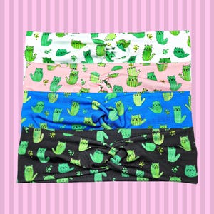 May include: Four headbands with a knotted design are displayed. Each headband features a repeating pattern of green cat-cactus illustrations and paw prints on different colored backgrounds: white, pink, blue, and black.
