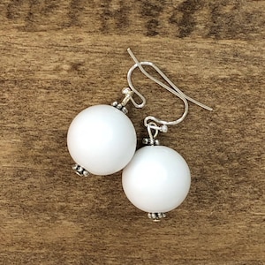 May include: A pair of white, round earrings with silver-colored hooks. The earrings have a simple design, with a small silver bead detail at the top and bottom of each white sphere. The earrings are displayed on a wooden surface.