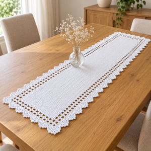 May include: A rectangular white crochet table runner featuring a dense central panel with a grid-like border and a scalloped edge. The runner is placed on a wooden surface with a clear glass vase containing dried white flowers centered on top.