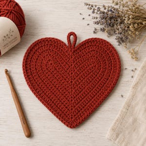 May include: A heart-shaped, dark red crocheted potholder with a small hanging loop at the top. Beside it lies a wooden crochet hook with 7.0 mm printed on the handle, a ball of dark red cotton yarn, and a small bunch of dried lavender flowers.