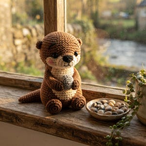 May include: A brown and cream-colored crocheted otter plush toy with black eyes and a small pink blush on its cheeks. The otter is sitting upright and holding a small, round, grey crocheted stone in its front paws, featuring a textured yarn finish.