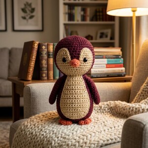 Könnte beinhalten: Ein handgefertigtes Häkel-Pinguin-Spielzeug mit einem dunkelburgunderfarbenen Körper, einem hellbeigen Bauch und Gesichtsfleck, zwei kleinen schwarzen Kunststoffaugen und einem kleinen orangefarbenen Schnabel. Die Plüschfigur steht auf zwei kleinen orangefarbenen Füßen.